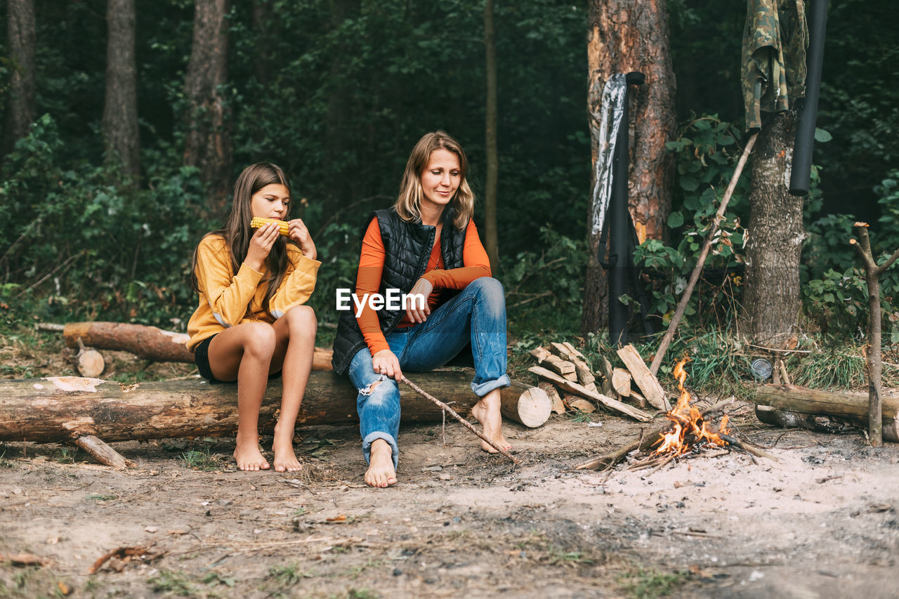 A mother with her teenage daughter is sitting on a tree near a campfire in the forest 