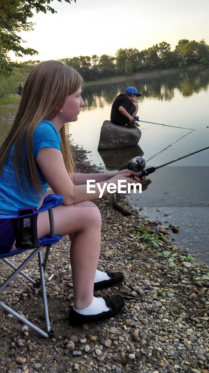 Siblings fishing on lakeshore against sky