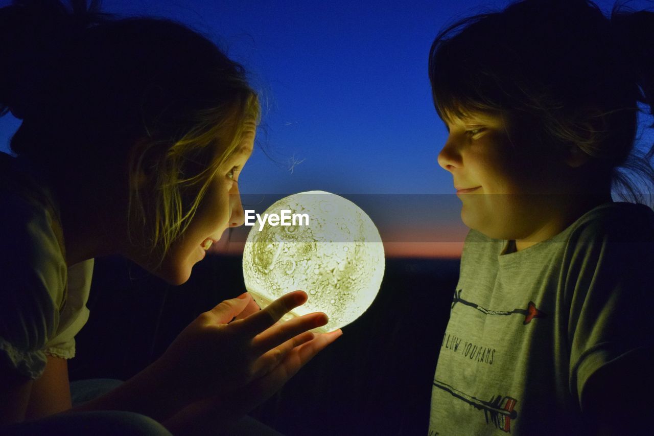 Side view of sisters playing with illuminated ball against sky during sunset