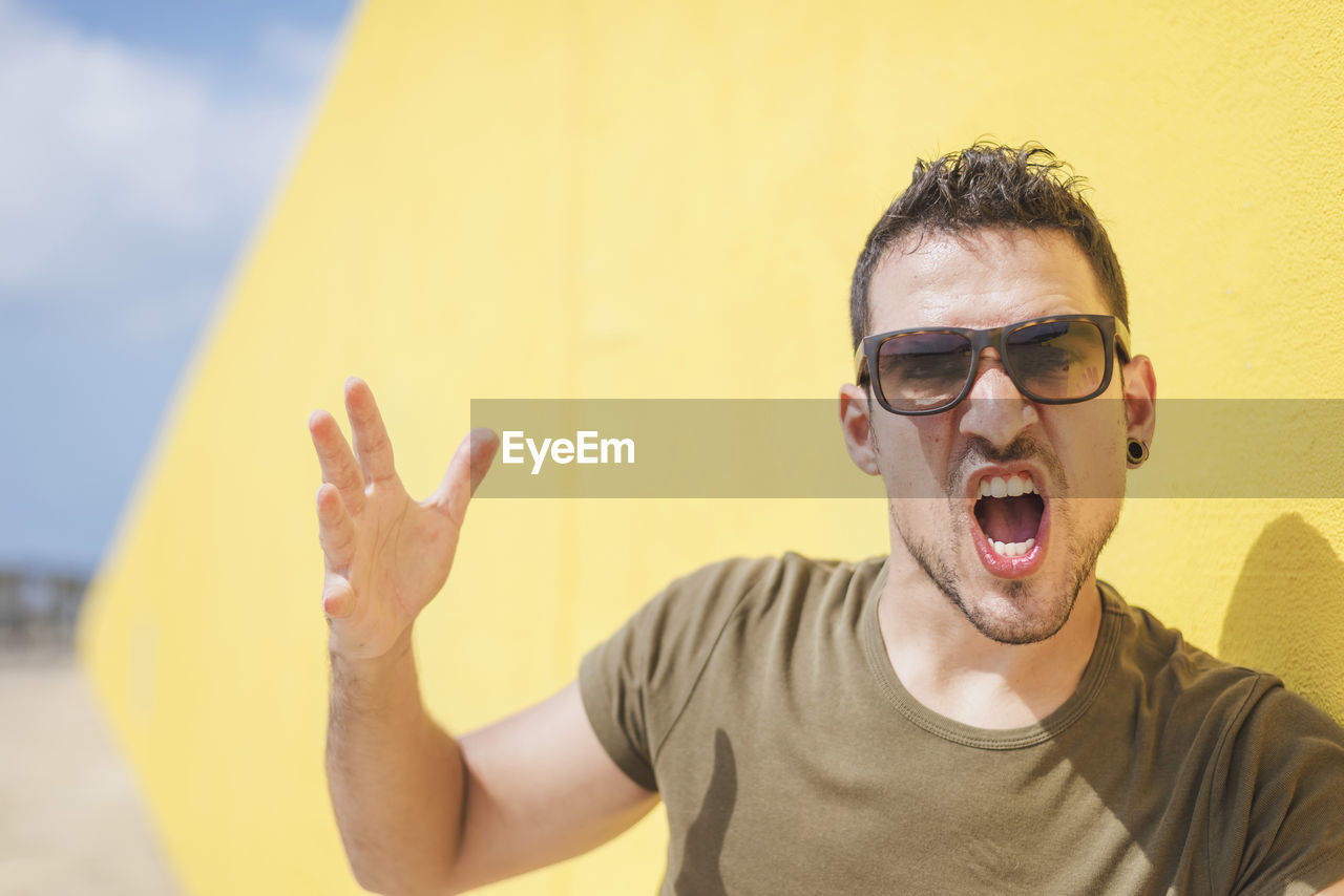 Portrait of man wearing sunglasses shouting while sanding against wall outdoors