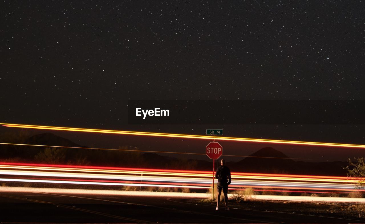 Man standing by light trails on road against sky at night