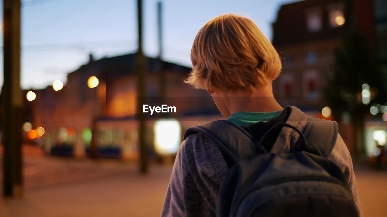 Rear view of boy standing with backpack in city at dusk