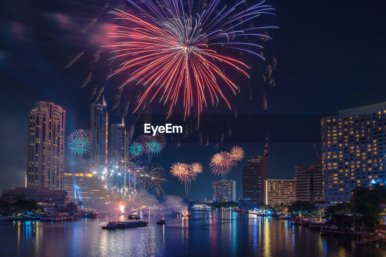 FIREWORK DISPLAY OVER RIVER BY BUILDINGS AGAINST SKY