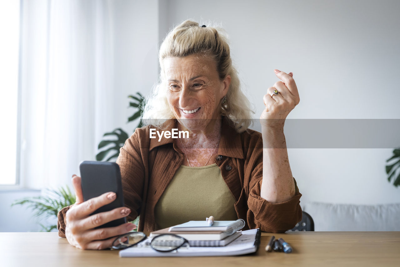 Happy senior businesswoman holding mobile phone at desk in home office