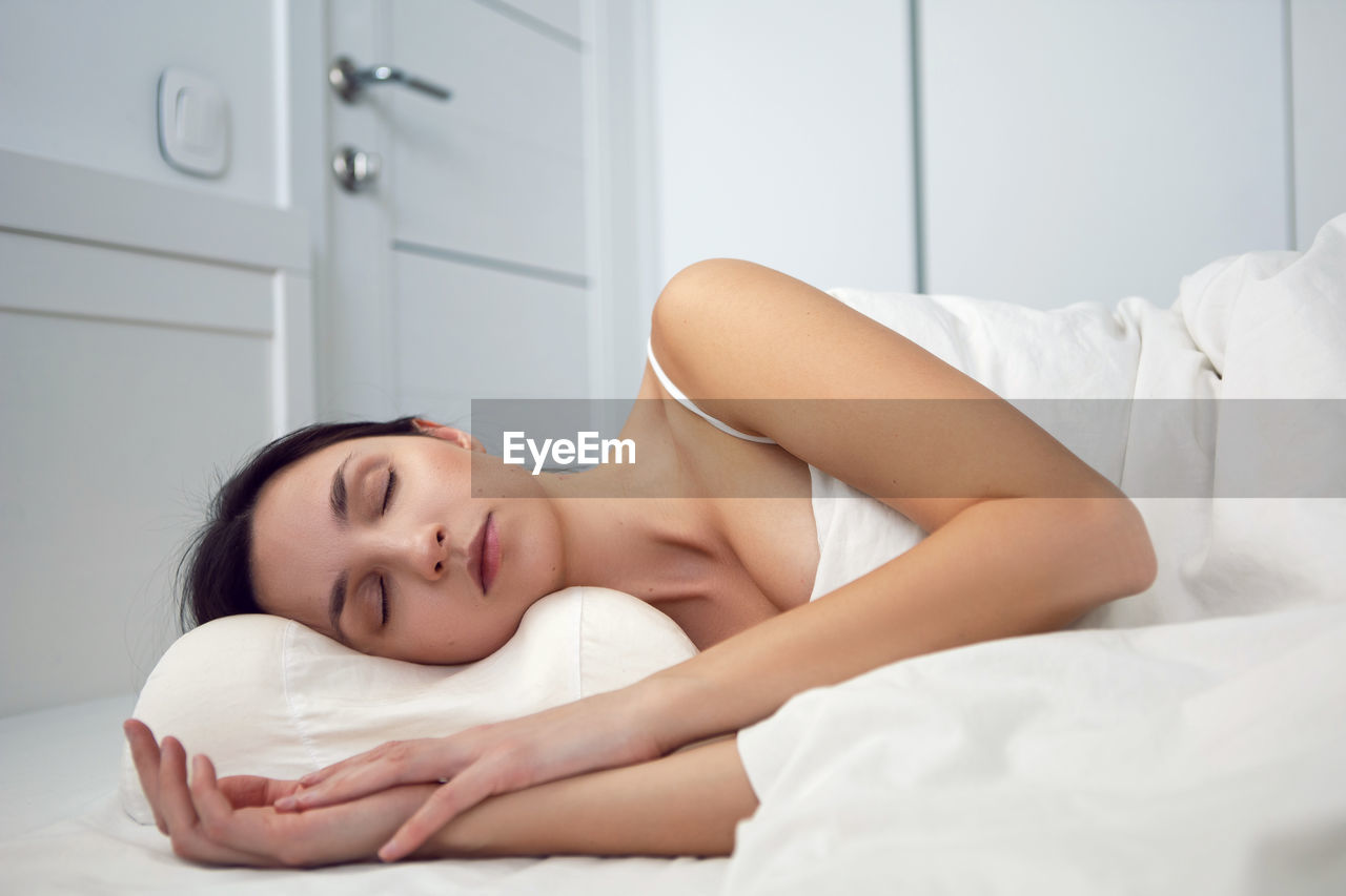 Woman in a white t-shirt is lying on the bed and a pillow is under the blanket in the room