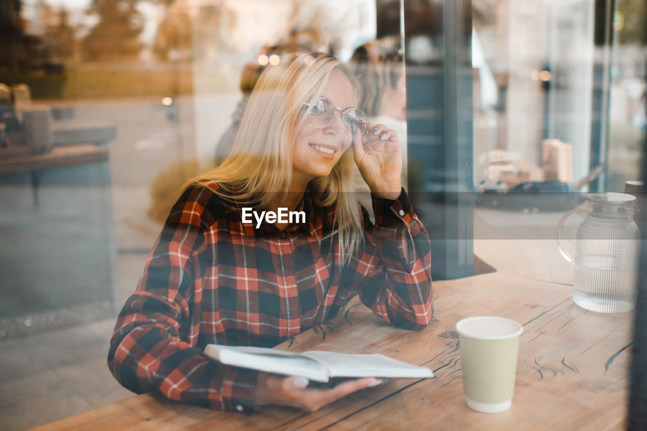 Smiling stylish young woman wear glasses and reading paper book in cafe through window close up.