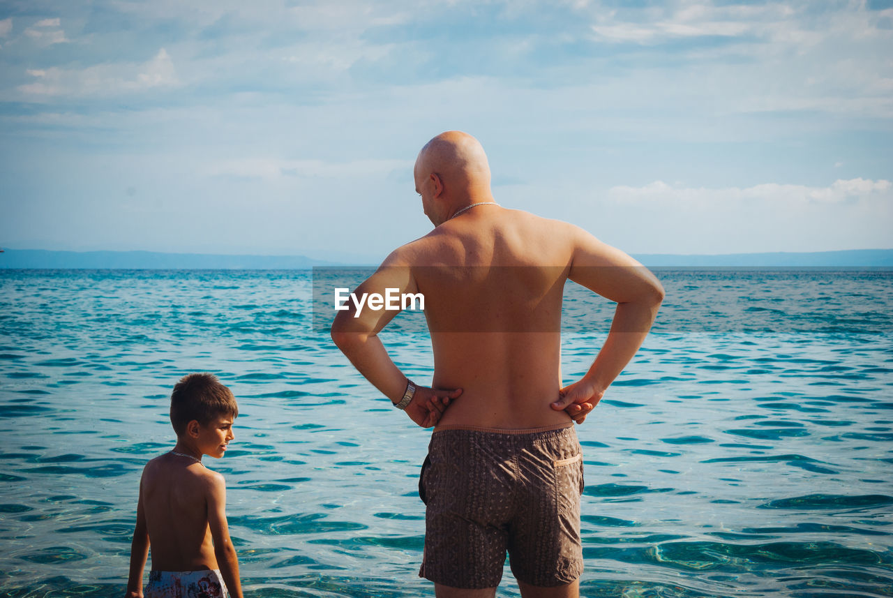 Rear view of shirtless father and son standing at beach against sky