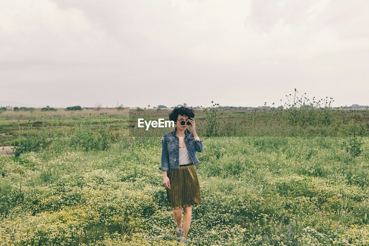 Portrait of mid adult woman wearing sunglasses standing on grassy field against sky