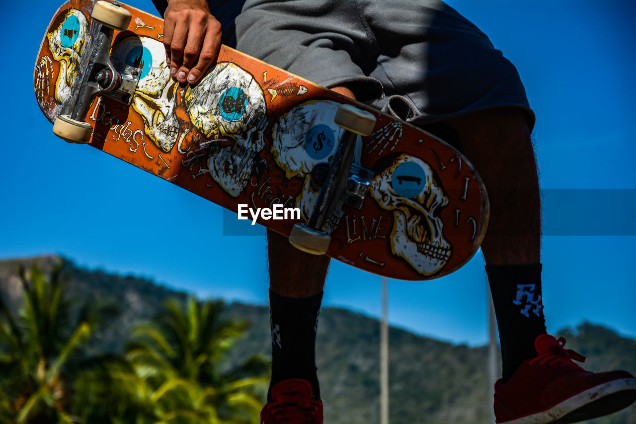 Low section of man holding skateboard against blue sky