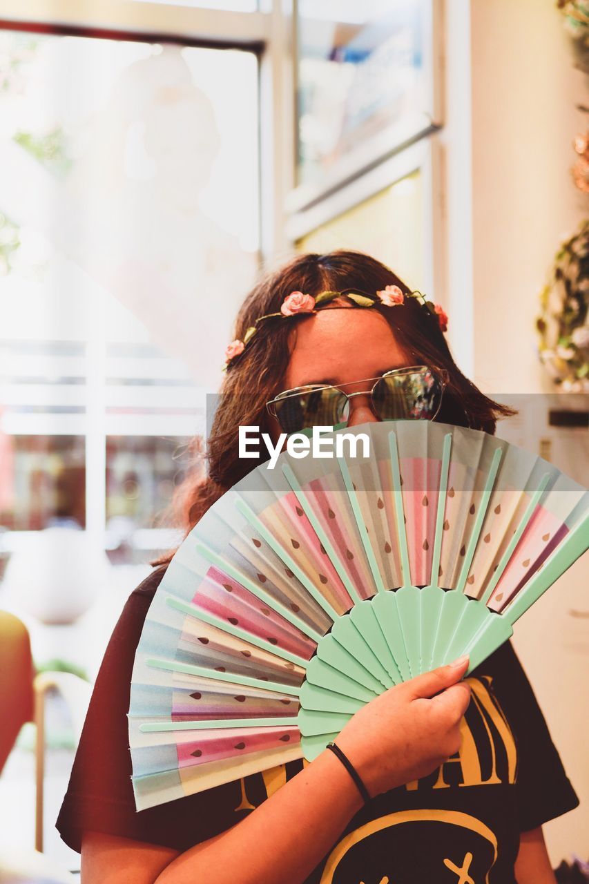 Portrait of woman in sunglasses covering face with hand fan at home