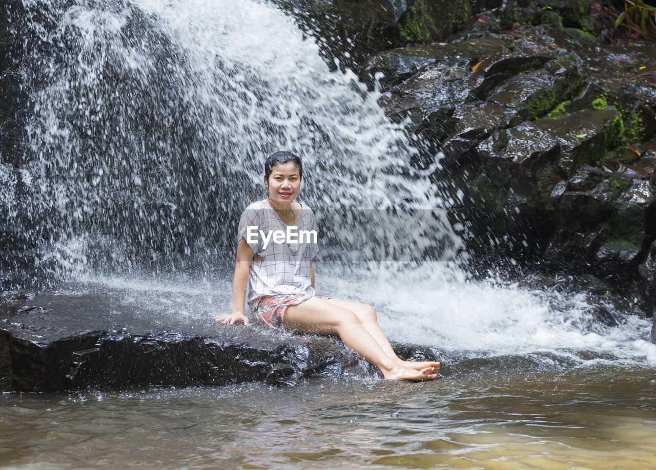 Portrait of smiling woman sitting on rock against waterfall