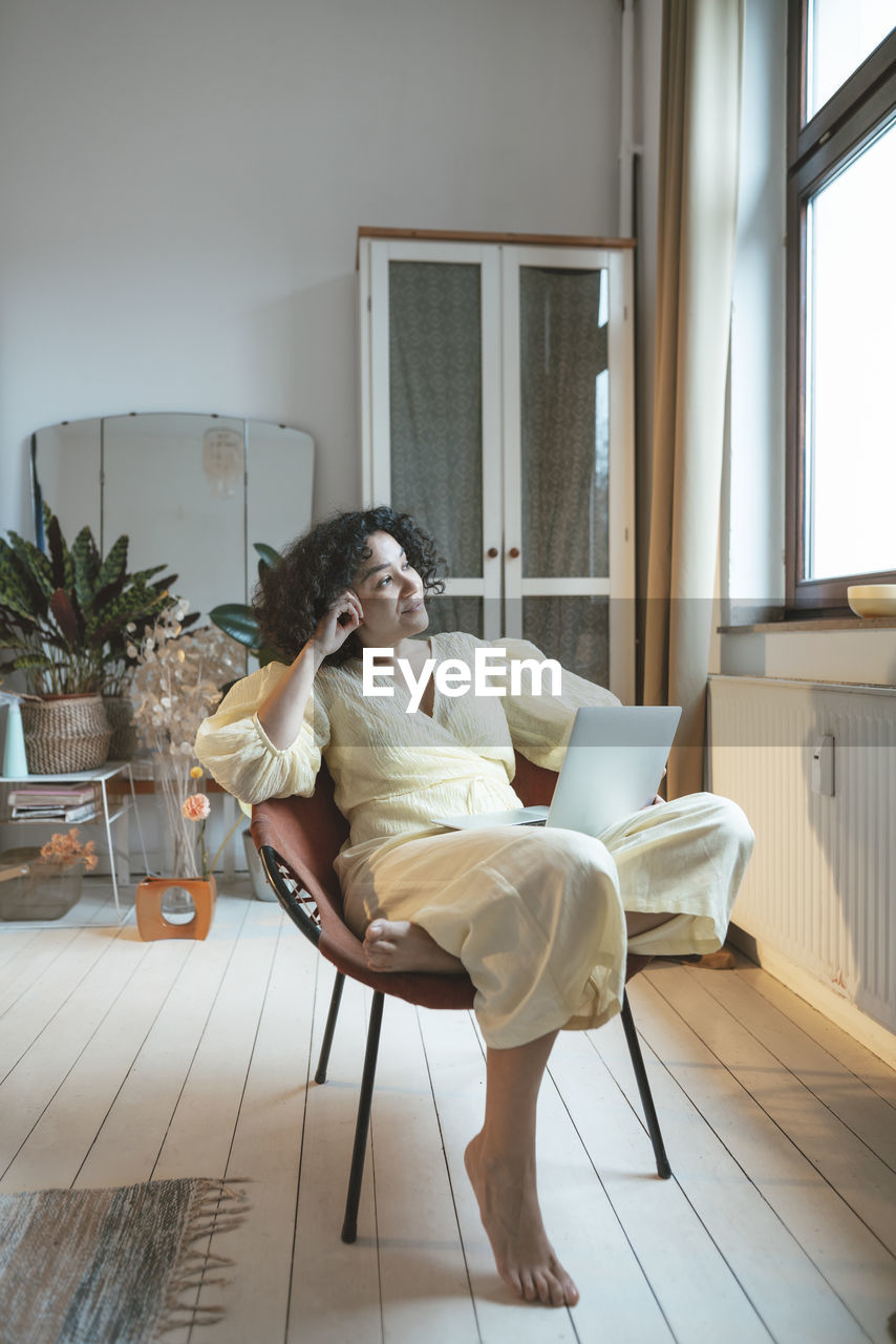 Smiling woman with laptop sitting with head in hands looking out through window at home