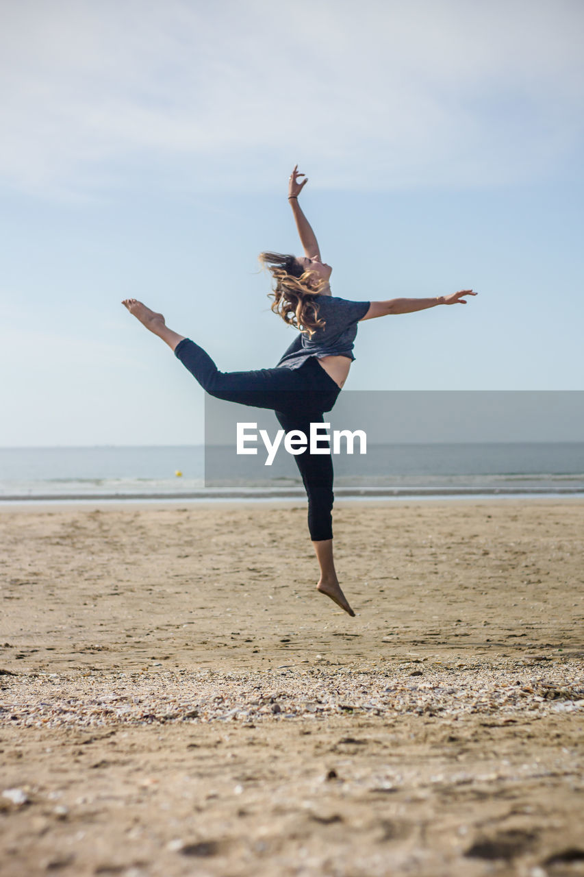 Side view of woman dancing on sand at beach against sky