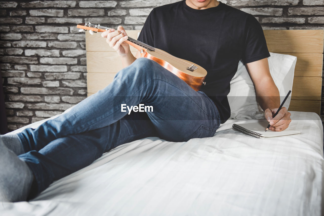 Low section of man playing guitar while writing on book at home