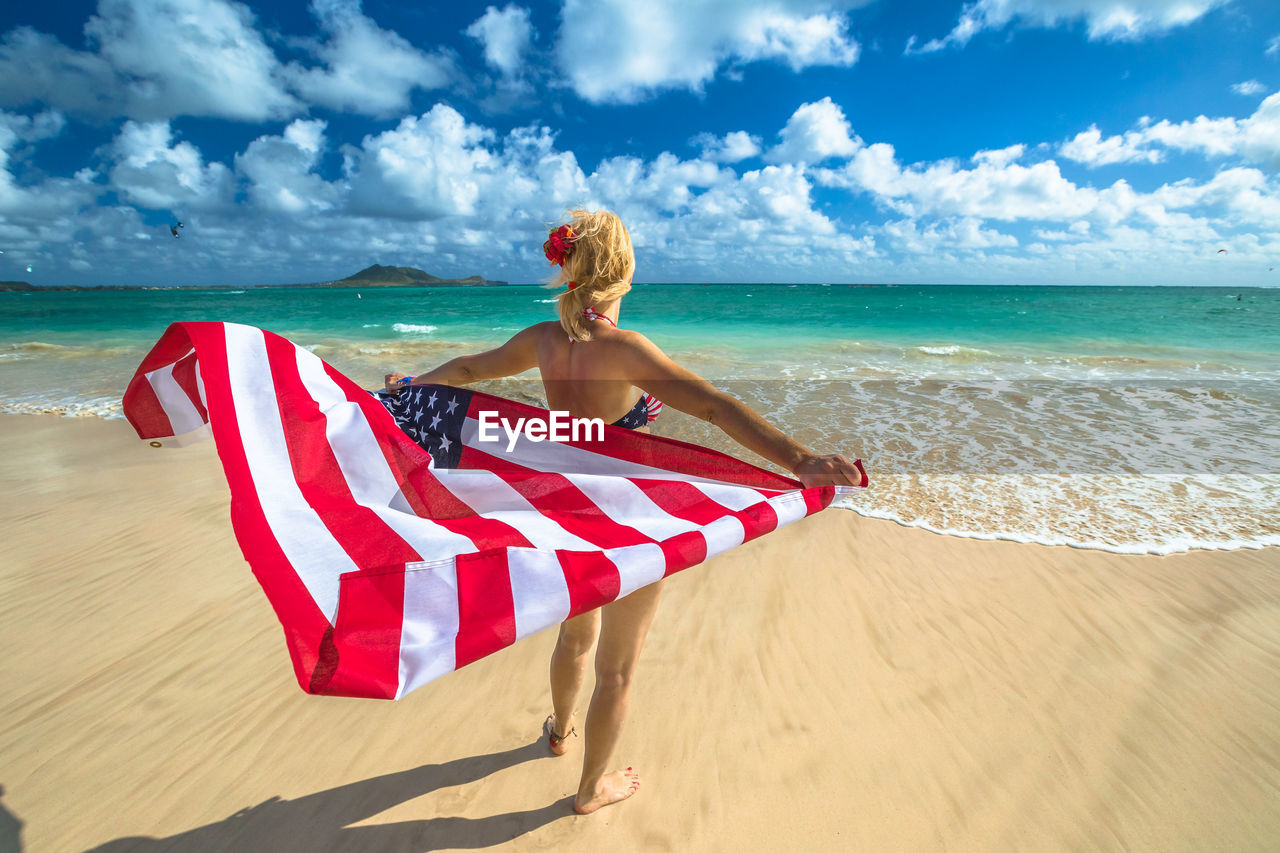 Rear view of woman holding american flag while standing at beach against blue sky