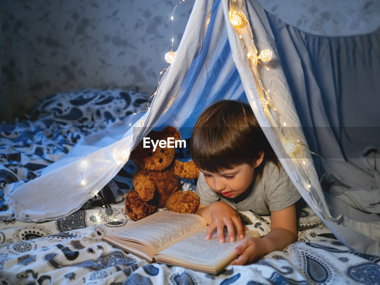 Little boy reads book. toddler plays in tent made of linen sheet on bed. 