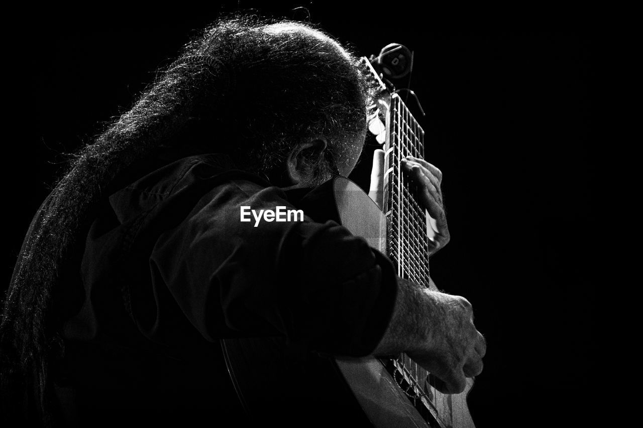 Side view of man playing guitar while sitting against black background