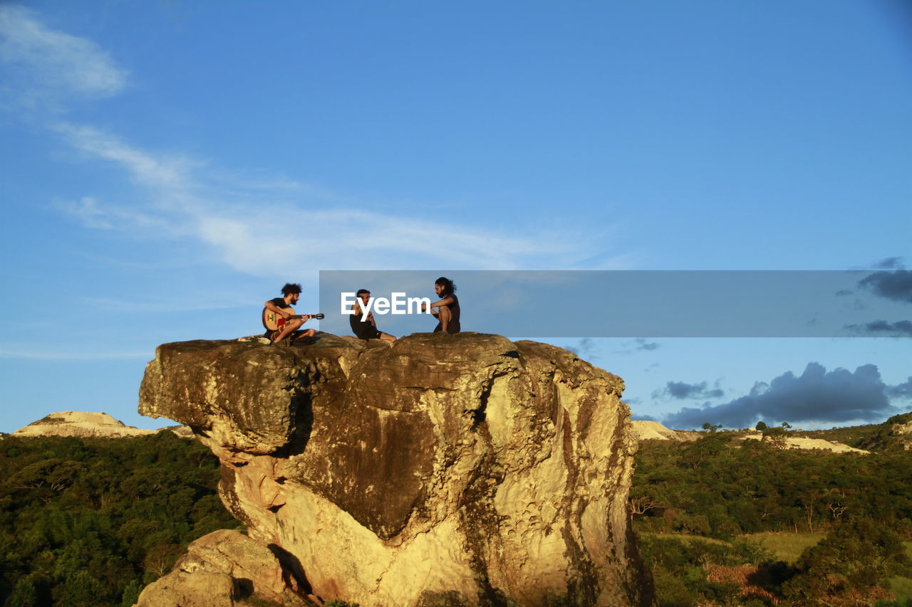 Friends sitting on rock formation against sky