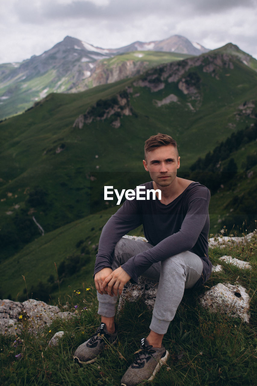 PORTRAIT OF YOUNG MAN SITTING ON MOUNTAINS AGAINST SKY