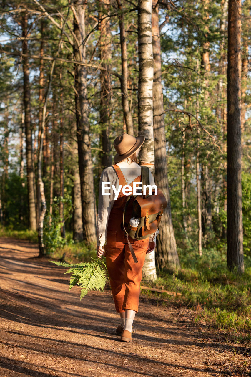 Woman botanist in orange overalls with backpack on ecological hiking trail in forest. 