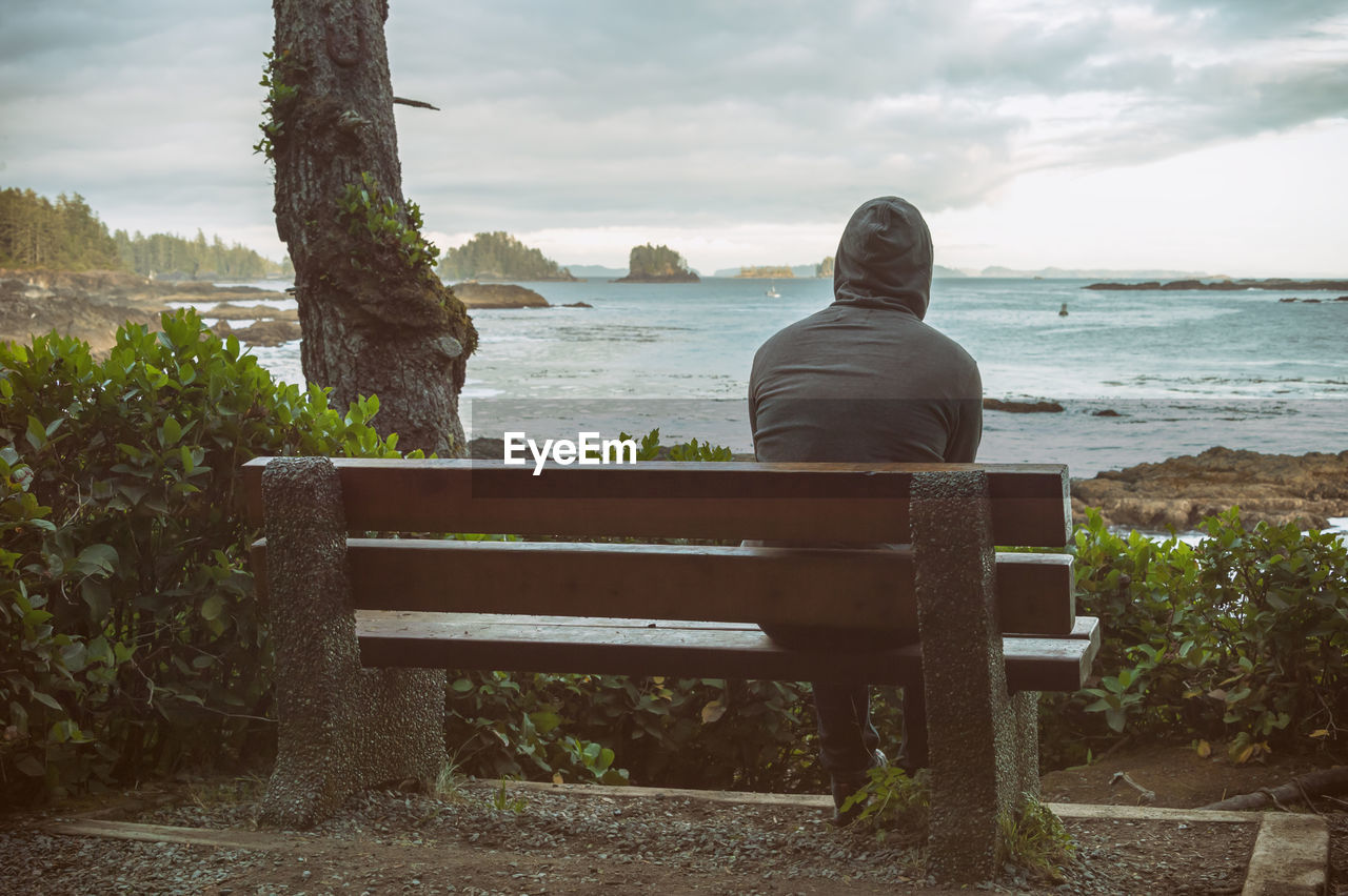 Rear view of man sitting on bench by sea against sky