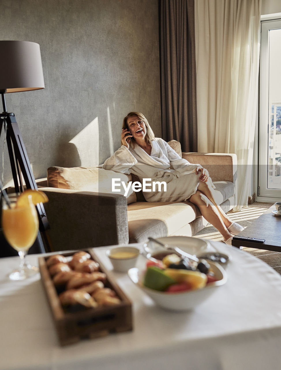 Fresh breakfast on table against senior woman talking through mobile phone while sitting on sofa at luxury hotel