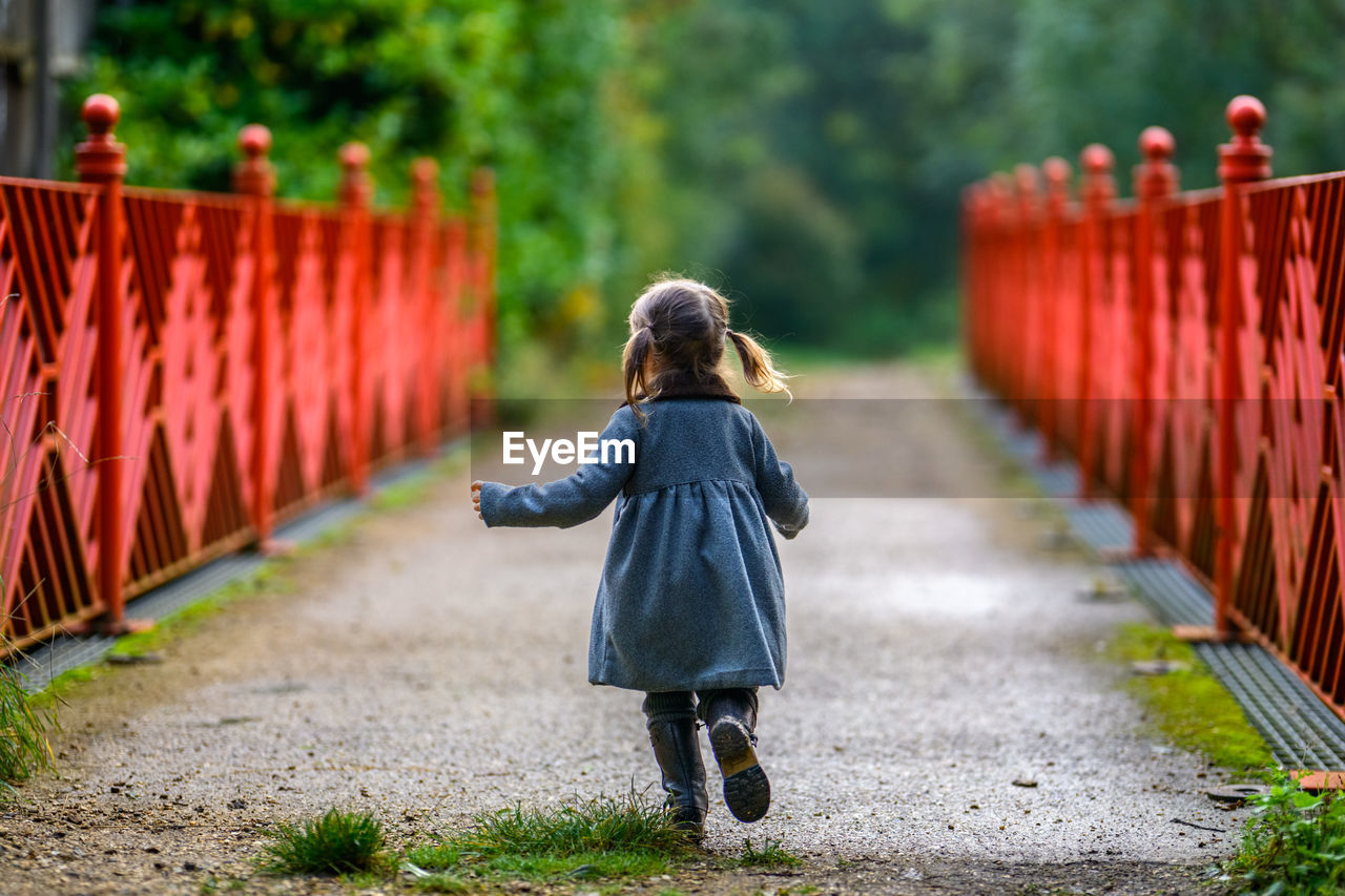 Little girl is running in the forest on a red bridge wearing coat and boots