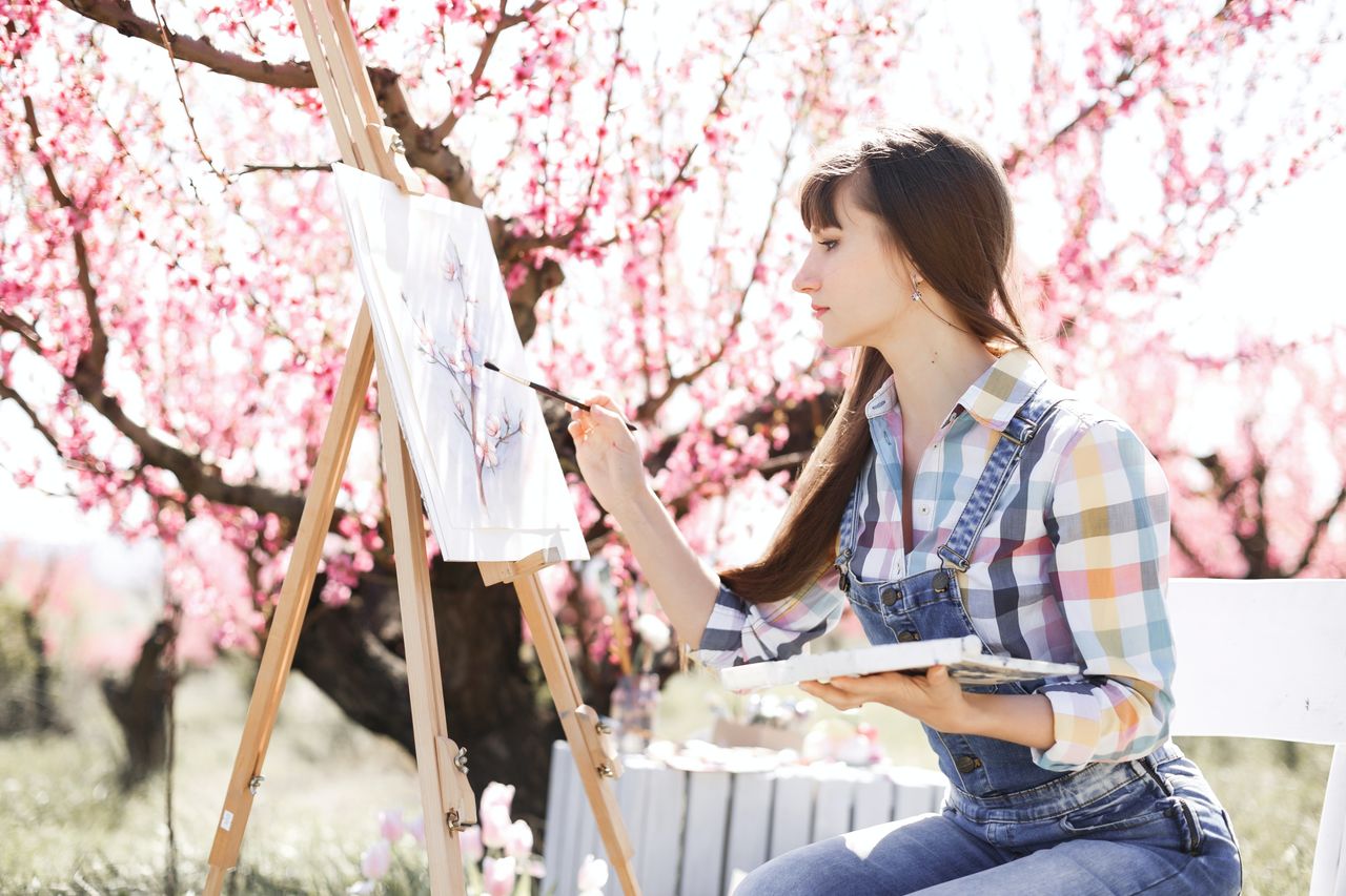 spring, plant, one person, adult, flower, women, young adult, flowering plant, blossom, nature, casual clothing, tree, springtime, pink, cherry blossom, three quarter length, beauty in nature, lifestyles, day, sitting, looking, hairstyle, freshness, outdoors, leisure activity, happiness, growth, holding, fragility, smiling, emotion, brown hair, side view, relaxation, sunlight, jeans, cherry tree