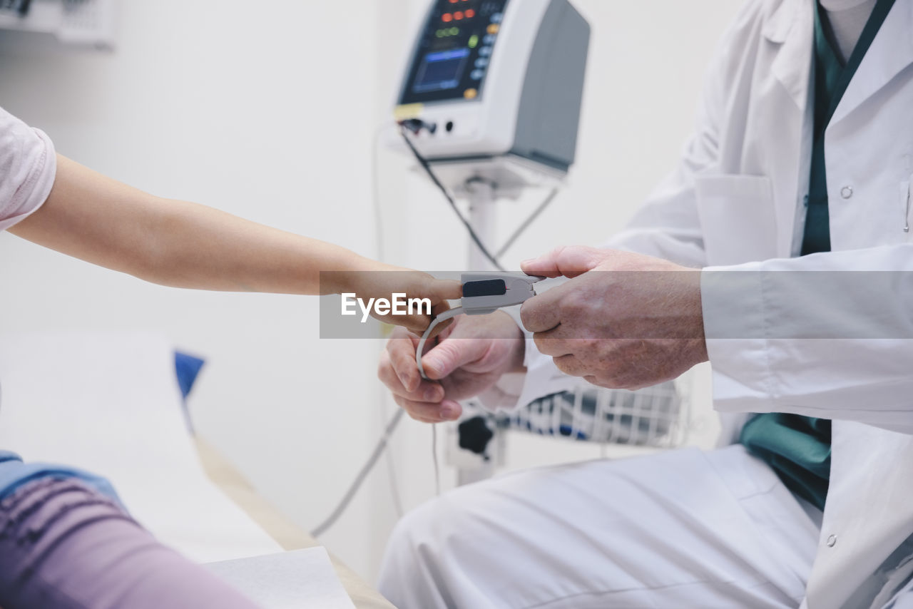 Midsection of mature doctor adjusting pulse oxymeter on girl's finger at hospital