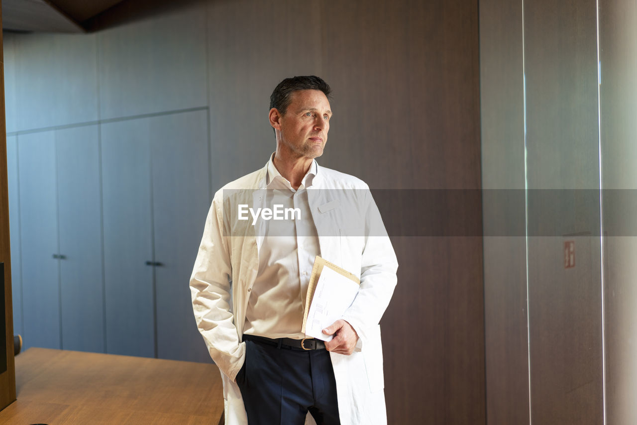 Thoughtful doctor wearing lab coat standing with hand in pocket at hospital