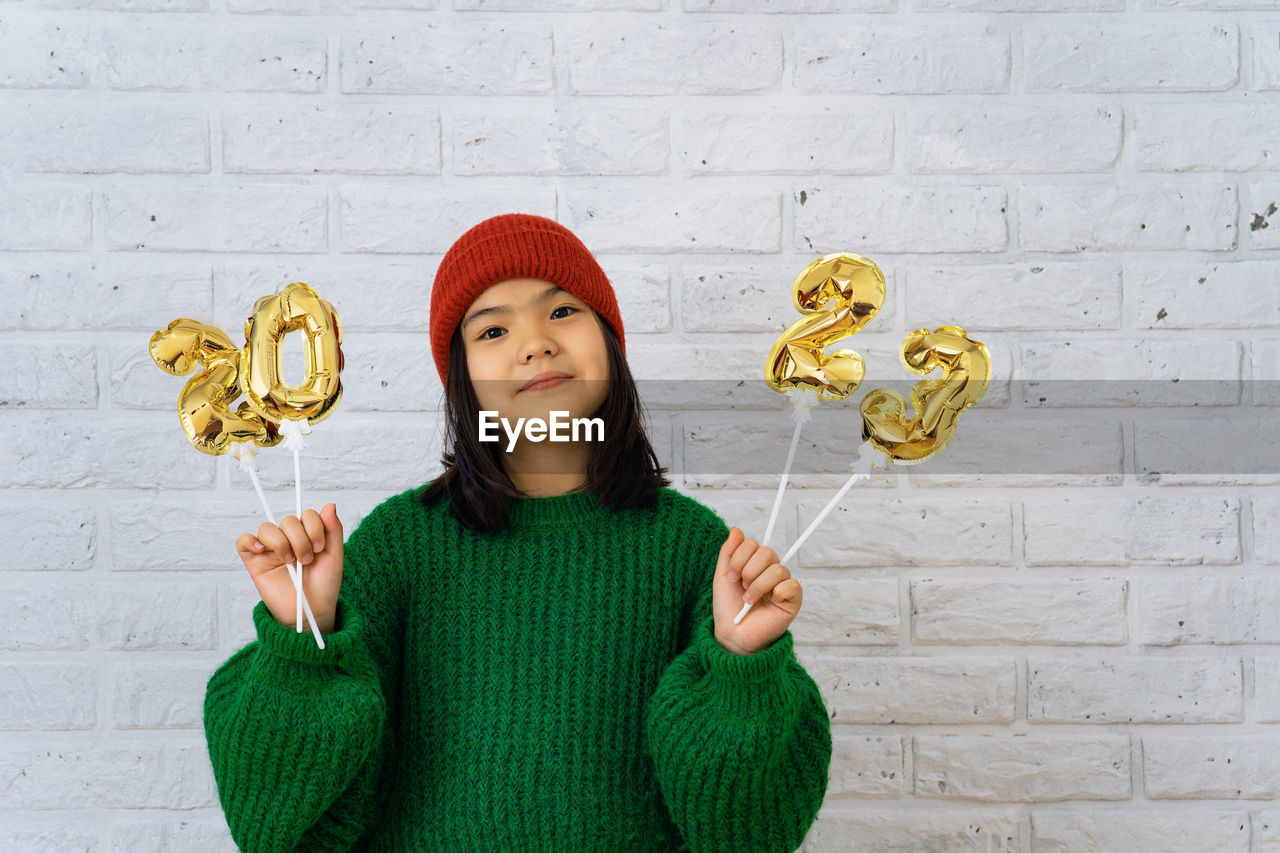 Asian cute girl in red cap and green sweater holding foil balloons with numbers 2023