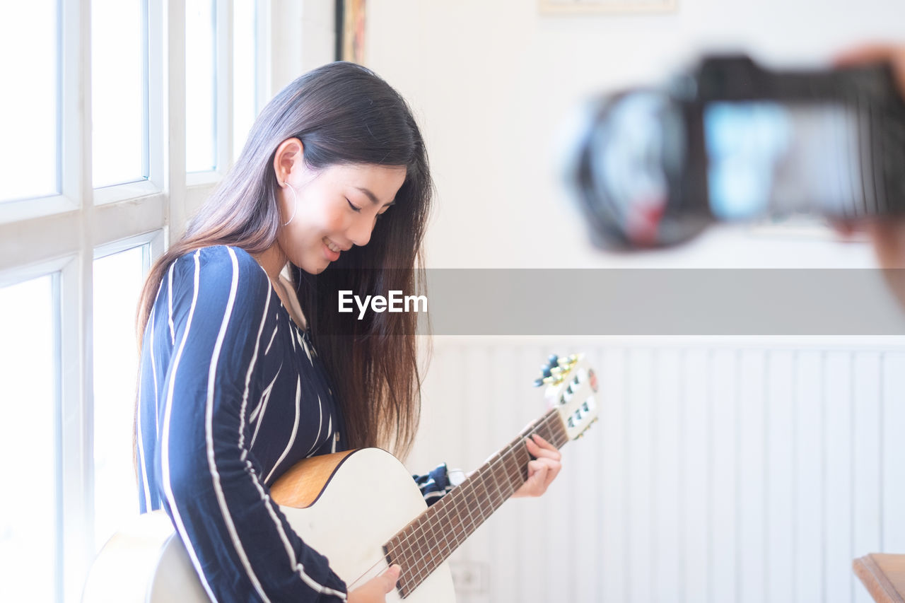 Smiling young woman blogging while playing guitar by window at home