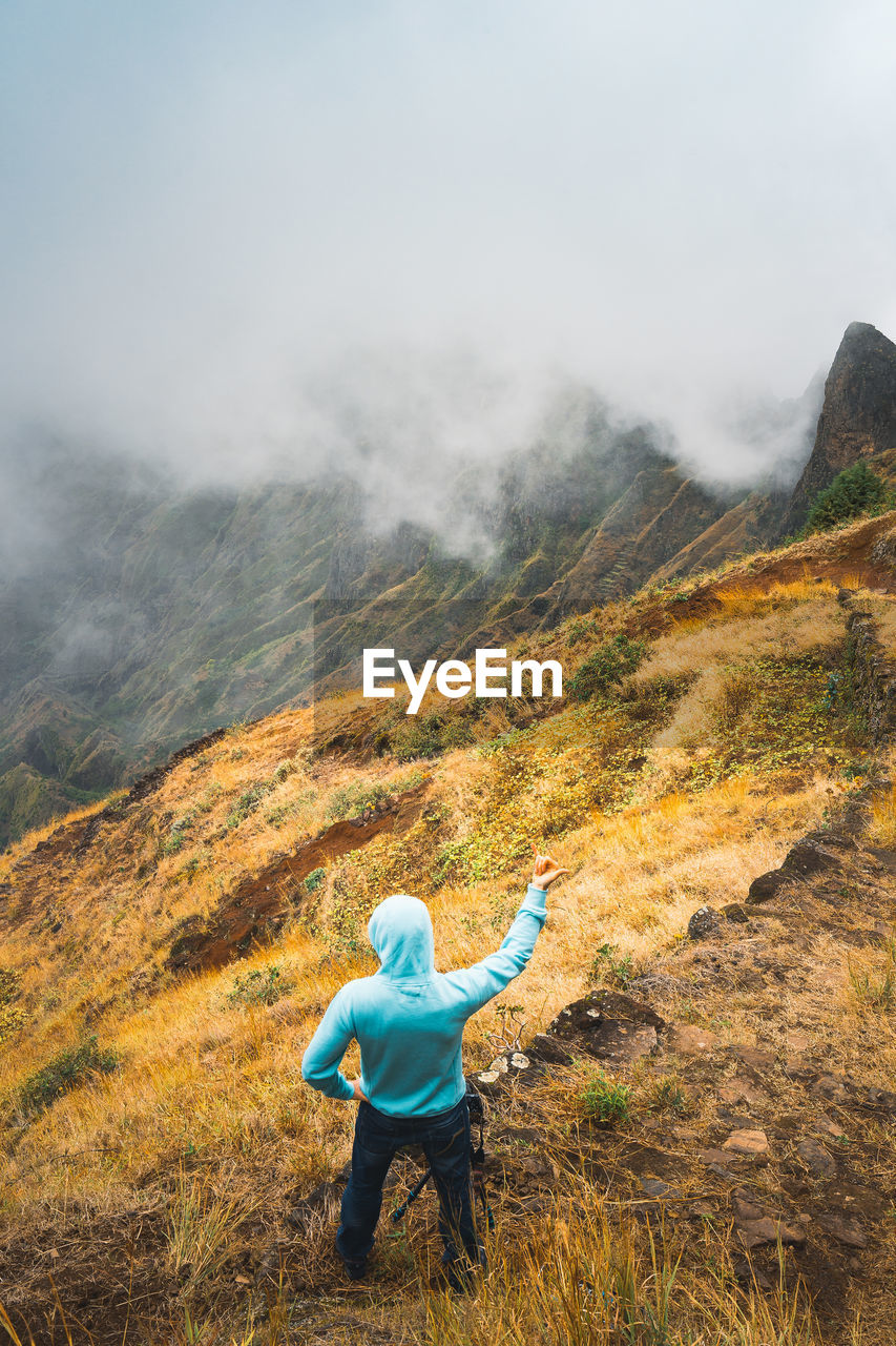 High angle view of man gesturing while standing on cliff during foggy mountain