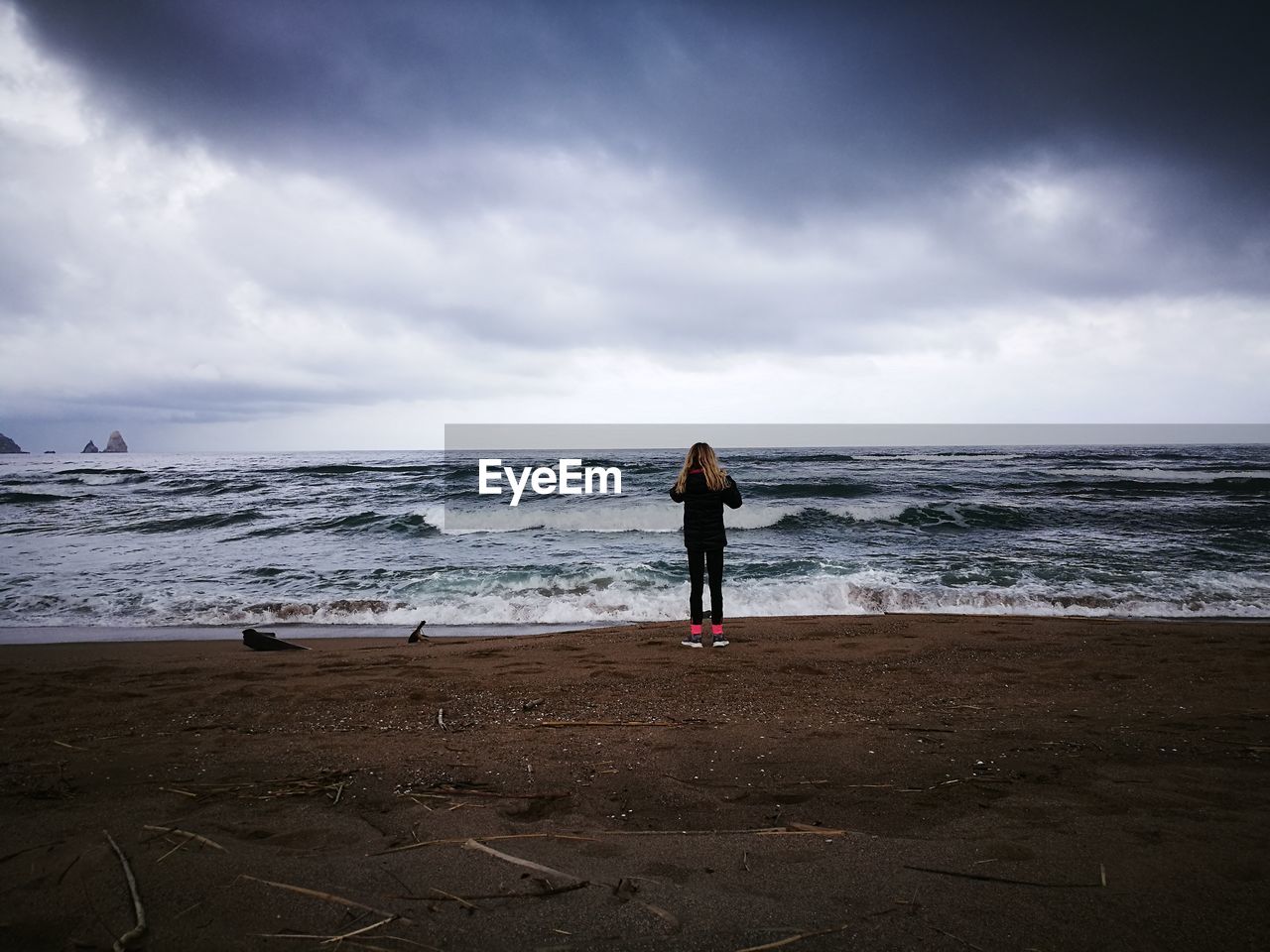 Rear view of woman standing at shore against cloudy sky