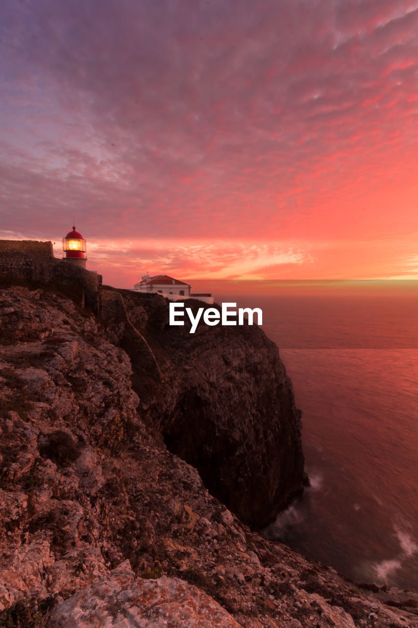 Coastline landscape at sunset with reddish sky and a lighthouse lit on the cape of san vicente, portugal