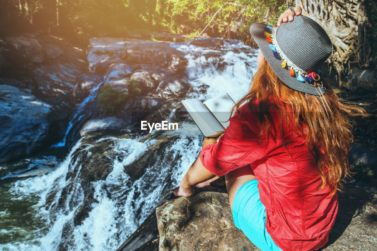 REAR VIEW OF WOMAN STANDING ON ROCK AGAINST WATERFALL