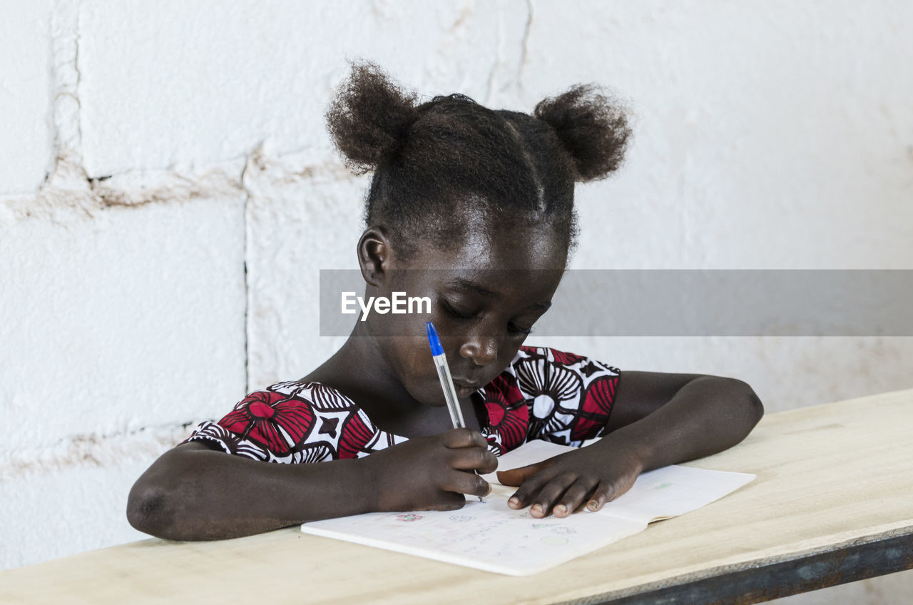 Girl writing while sitting at table against wall