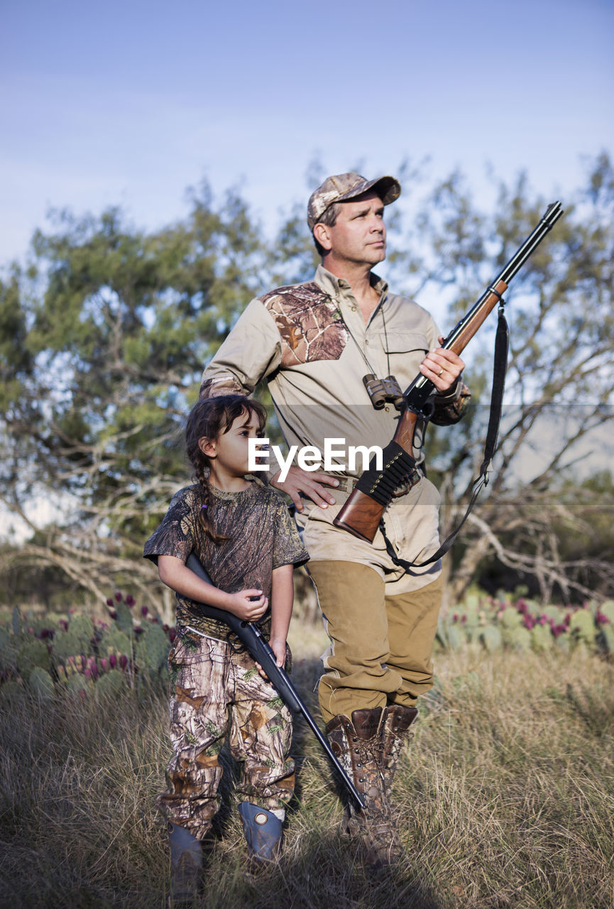 Hunters holding rifle and looking away while standing on grassy field