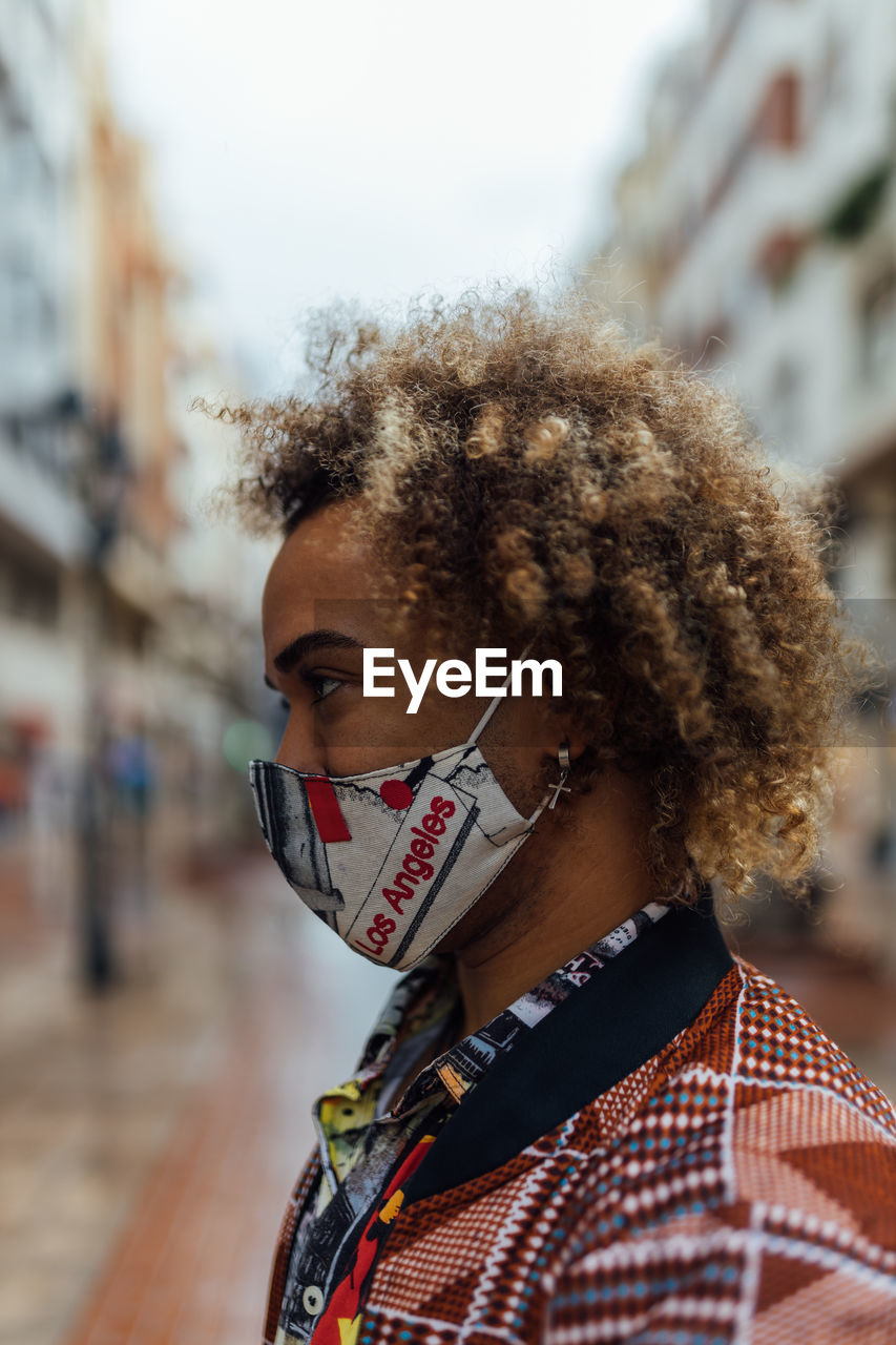 Side view of modern stylish african american male with curly hair dressed in trendy colorful informal clothes and protective mask standing against blurred urban environment and looking away