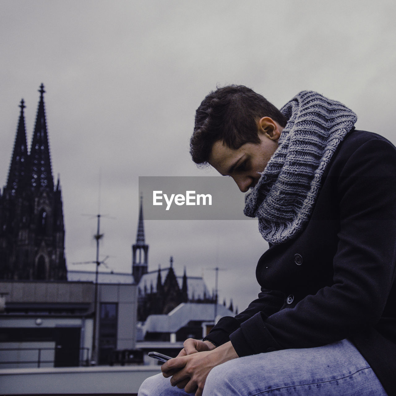 Side view of young man using mobile phone while sitting at building terrace with cologne cathedral in background