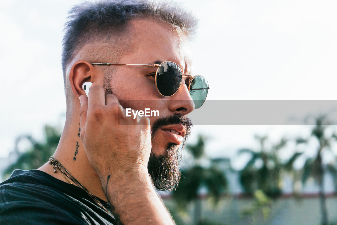 Side view of thoughtful bearded male with tattoos in casual clothes and sunglasses wearing earphones and looking away on street against green trees in sunny day