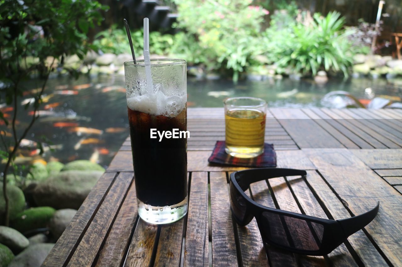 Close-up of drinks and sunglasses on wooden table against lake in forest