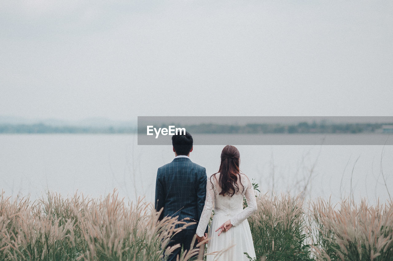 REAR VIEW OF COUPLE STANDING ON SHORE AGAINST SKY