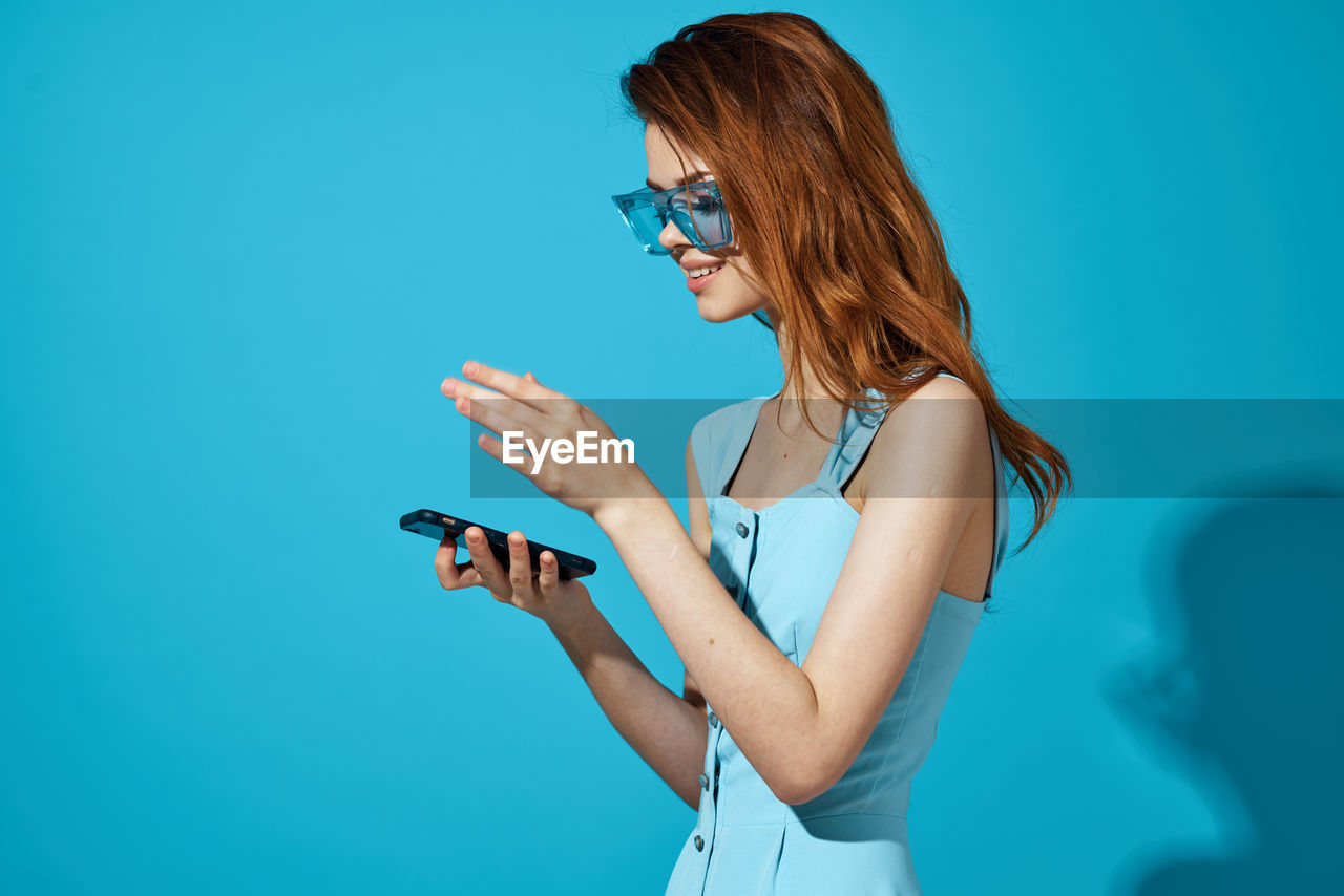 Young woman using phone while standing against blue sky