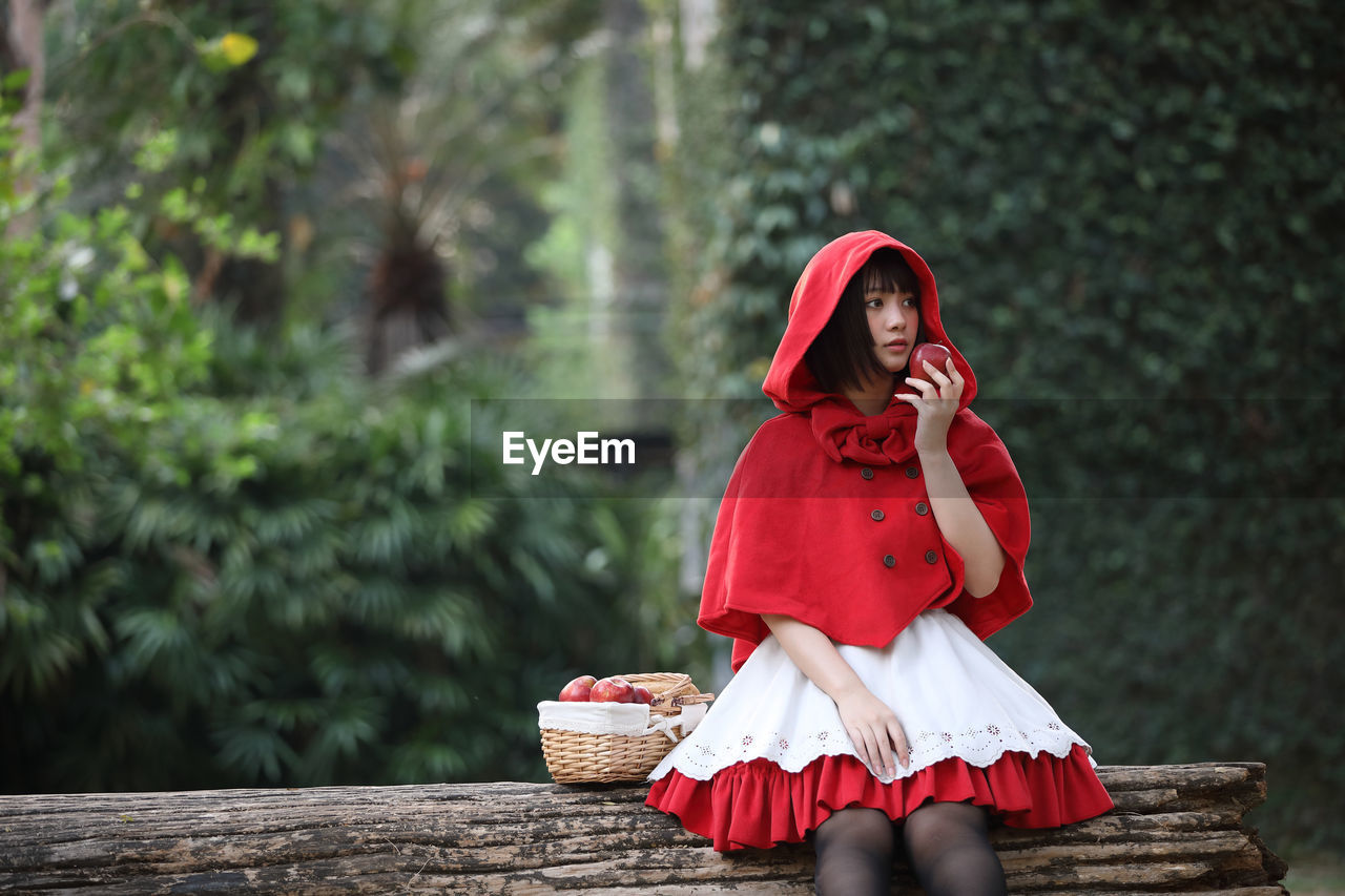 Young woman with fruits in basket looking away while sitting against tree