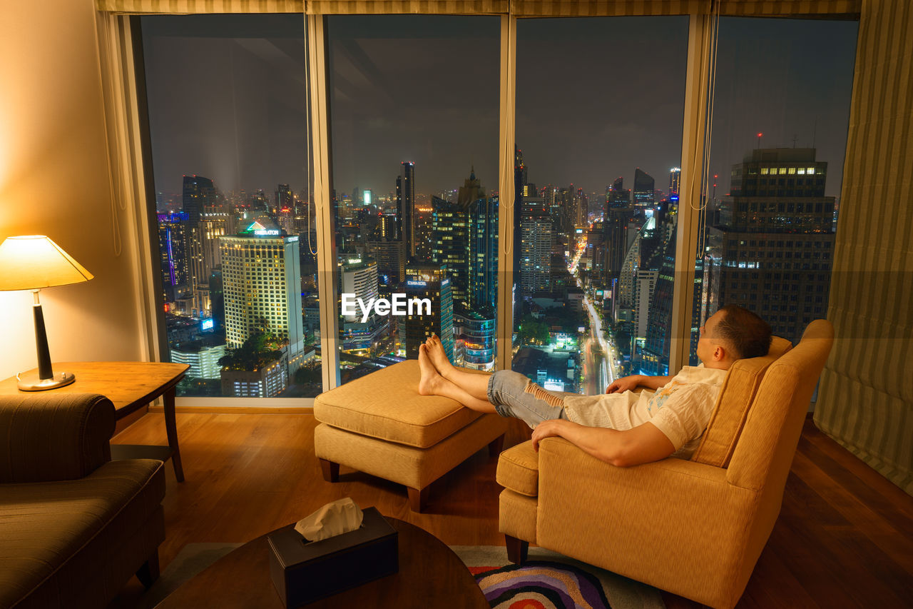 Man resting on chair in cozy room interior in the evening and looks through window on night city