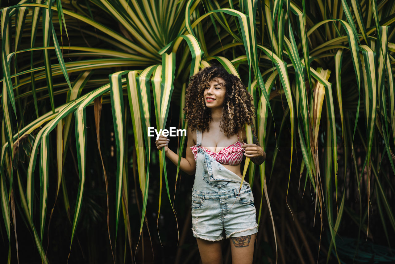 Seductive woman with curly hair standing against plants