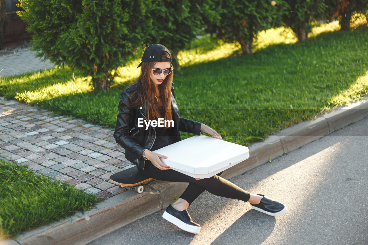 Young woman with pizza box sitting on skateboard against plants