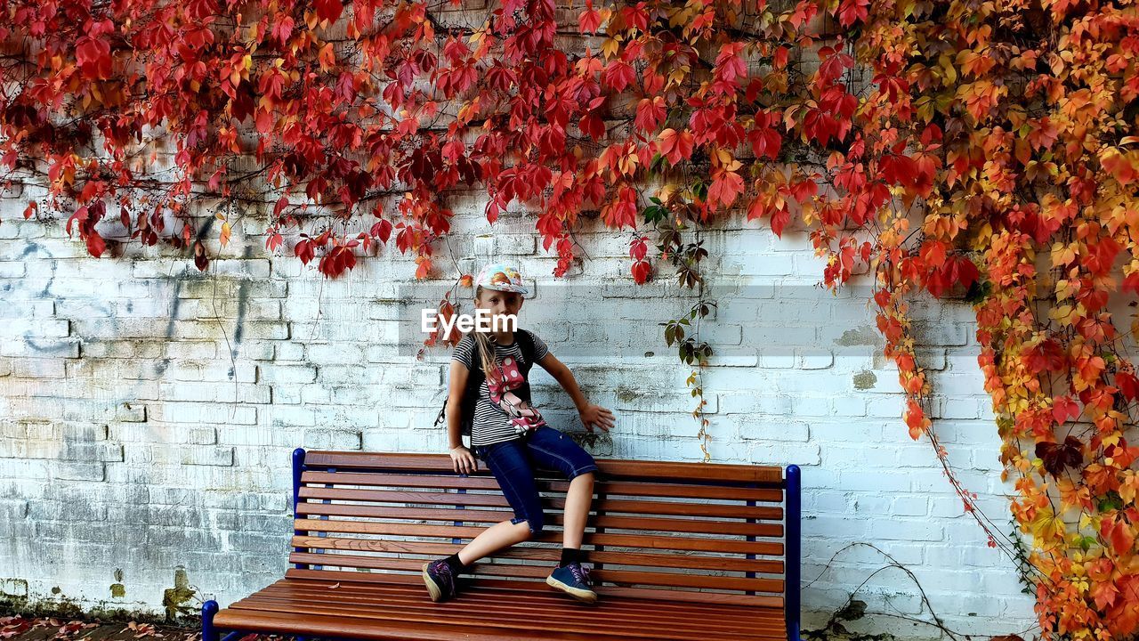 Portrait of smiling girl sitting on bench against wall during autumn
