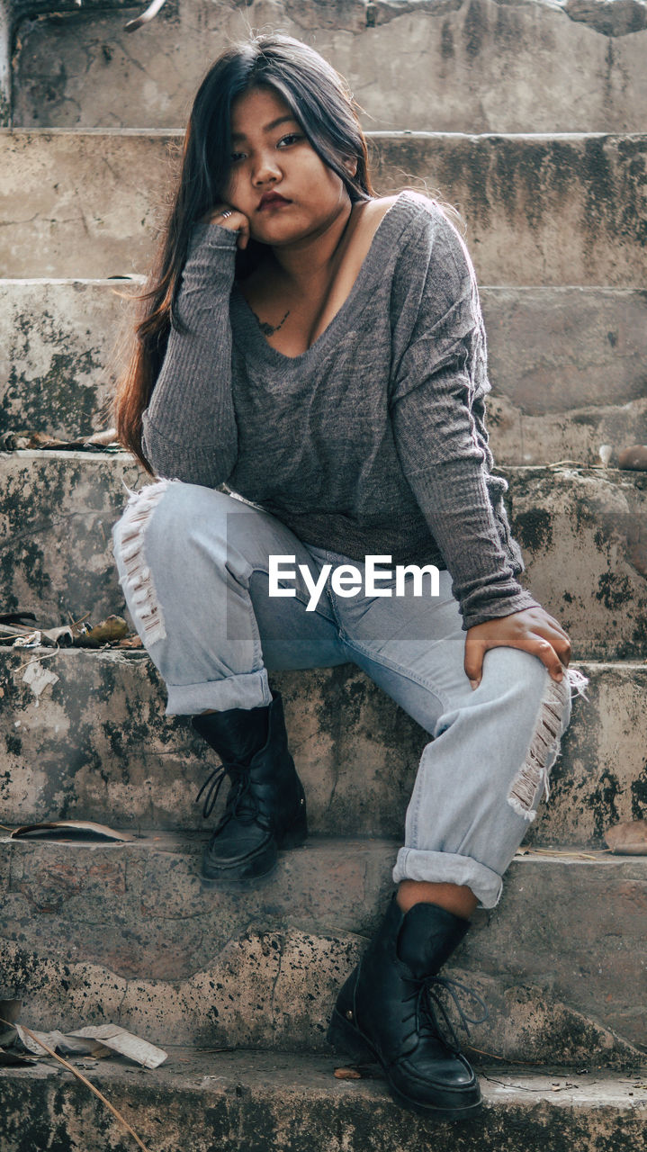 Young woman looking down while sitting against brick wall